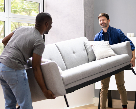 Two smiling men moving a sofa into a living room.