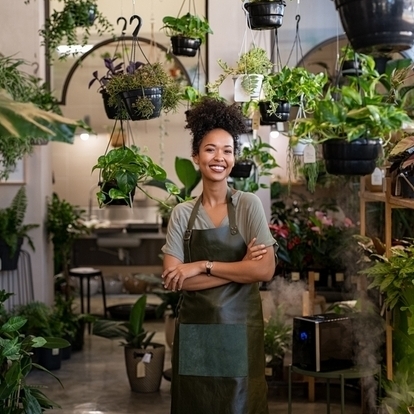 A smiling woman, the owner of a plant store, stands surrounded by potted plants.