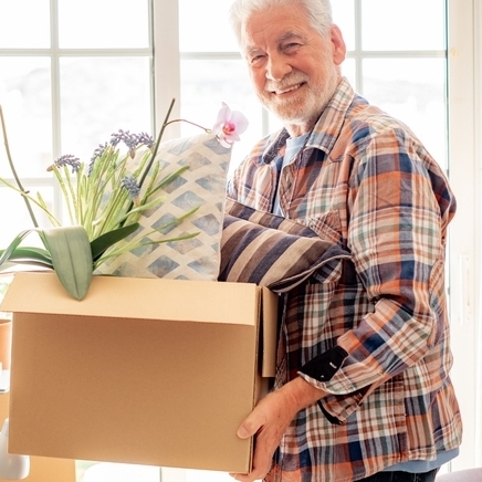 A smiling older man moving a cardboard box into a new home.