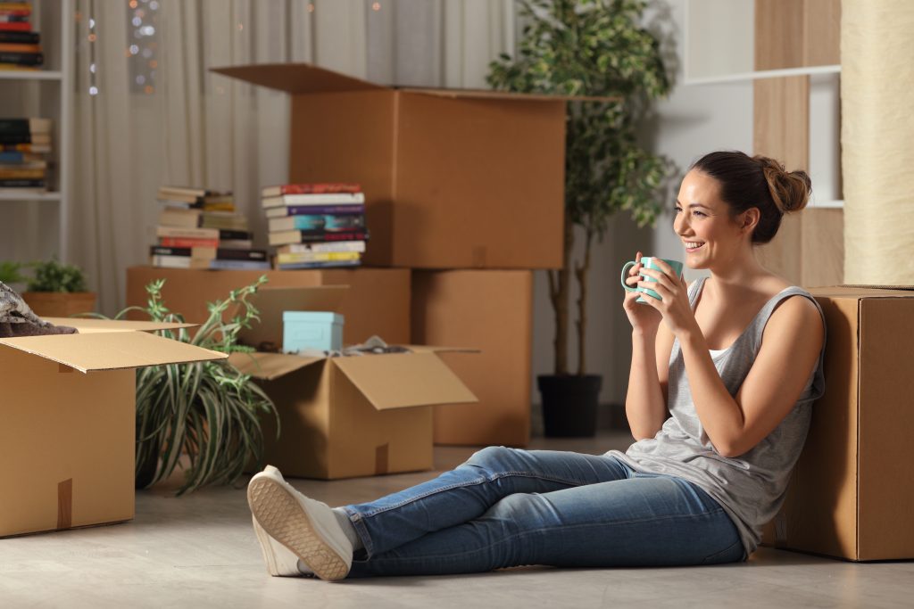 A happy woman sitting on the floor of her new apartment, surrounded by moving boxes.