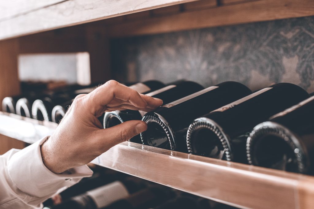 A hand reaching for a bottle of wine on a shelf filled with wine bottles.