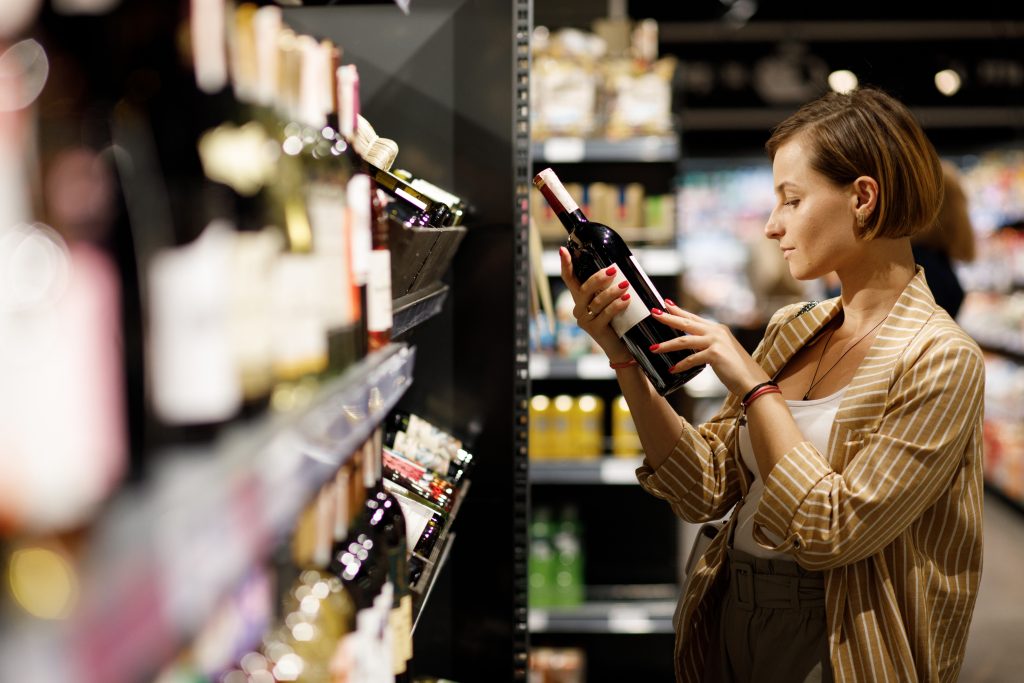 A woman looking at a bottle of wine in a liquor store aisle.