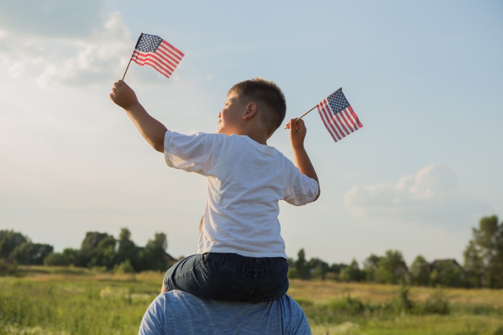 A little boy sitting on his dad's shoulders holding up two miniature American flags. 