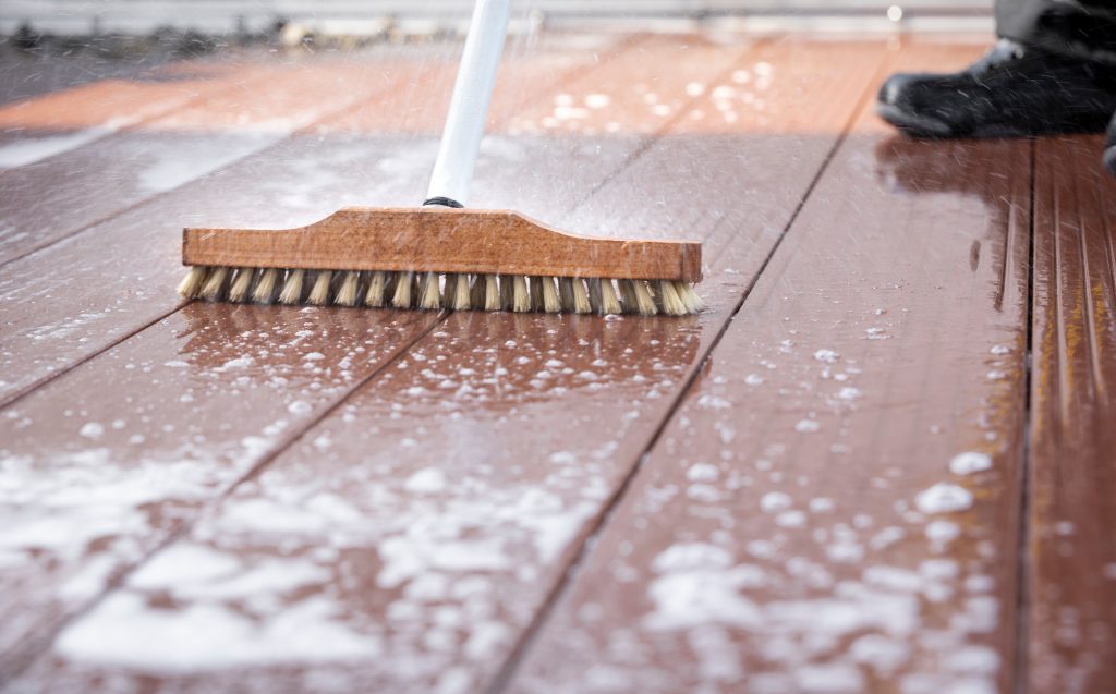 A heavy duty broom being used to scrub a wood deck with water and soap.