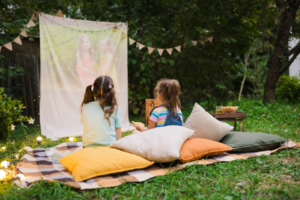Two young girls sitting outside on a blanket in front of a projector screen.