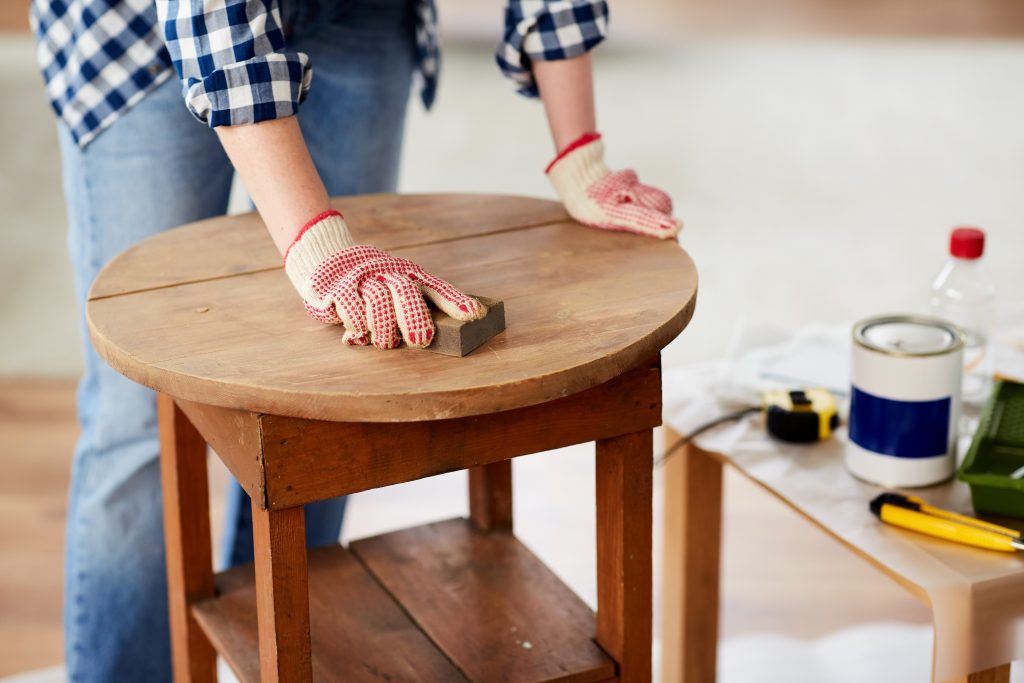 woman in work gloves sanding the top of a small table, with various tools and paint nearby.