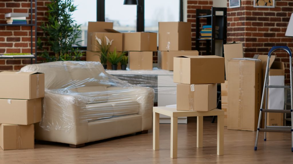 A room full of carboard boxes next to a large armchair wrapped in plastic covering for storage.
