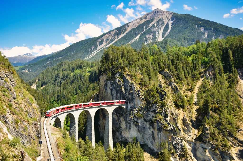 A mountainous aerial shot with a train moving along elevated tracks through the mountains.
