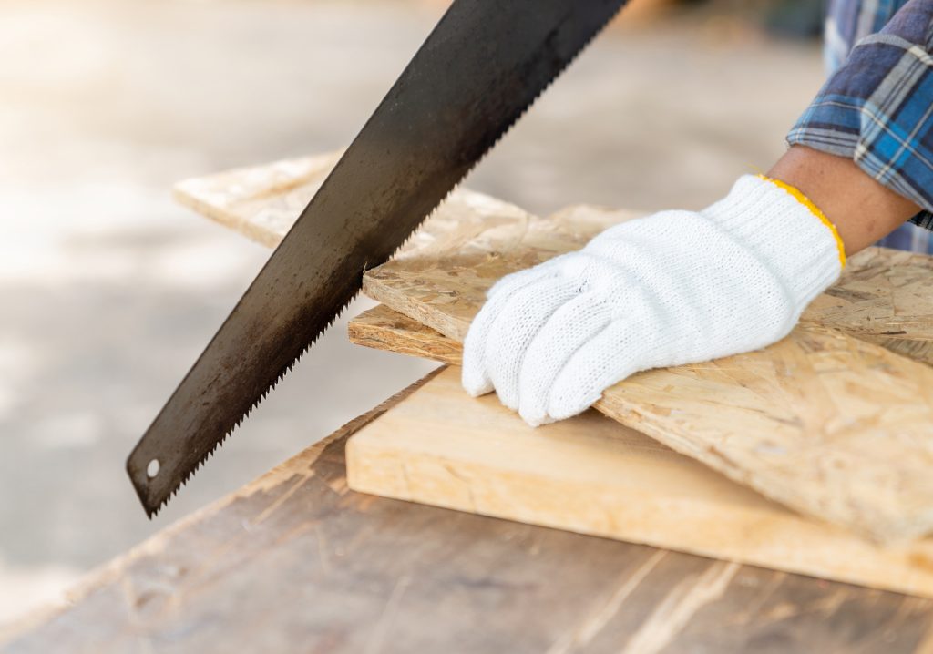 A man wearing work gloves sawing a piece of plywood.
