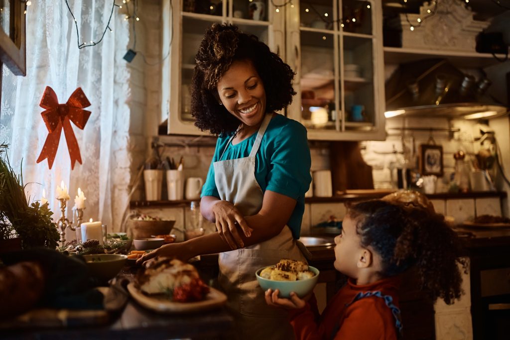 A woman cooking in a kitchen, looking down at a small child near the counter.