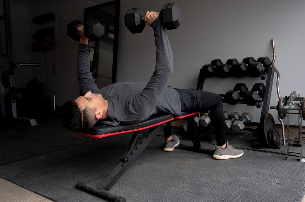 A man on his back on a weight bench, lifting dumbbells overhead.