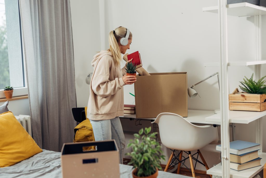 A college student unpacking in a dorm with a small bed and desk.