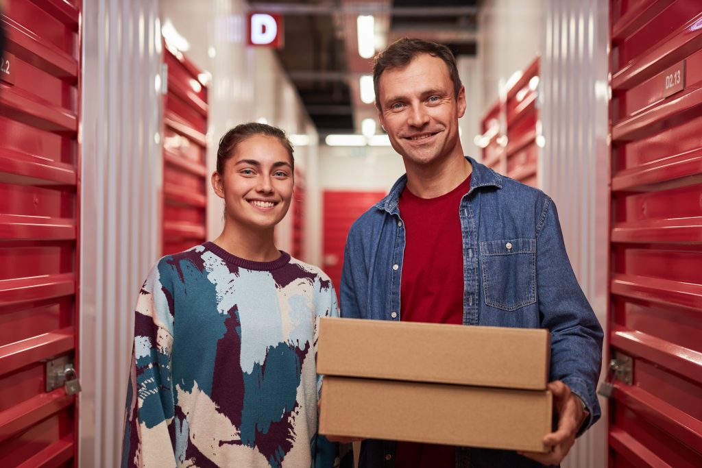 A father and daughter standing in a self storage corridor with a cardboard box in hand.