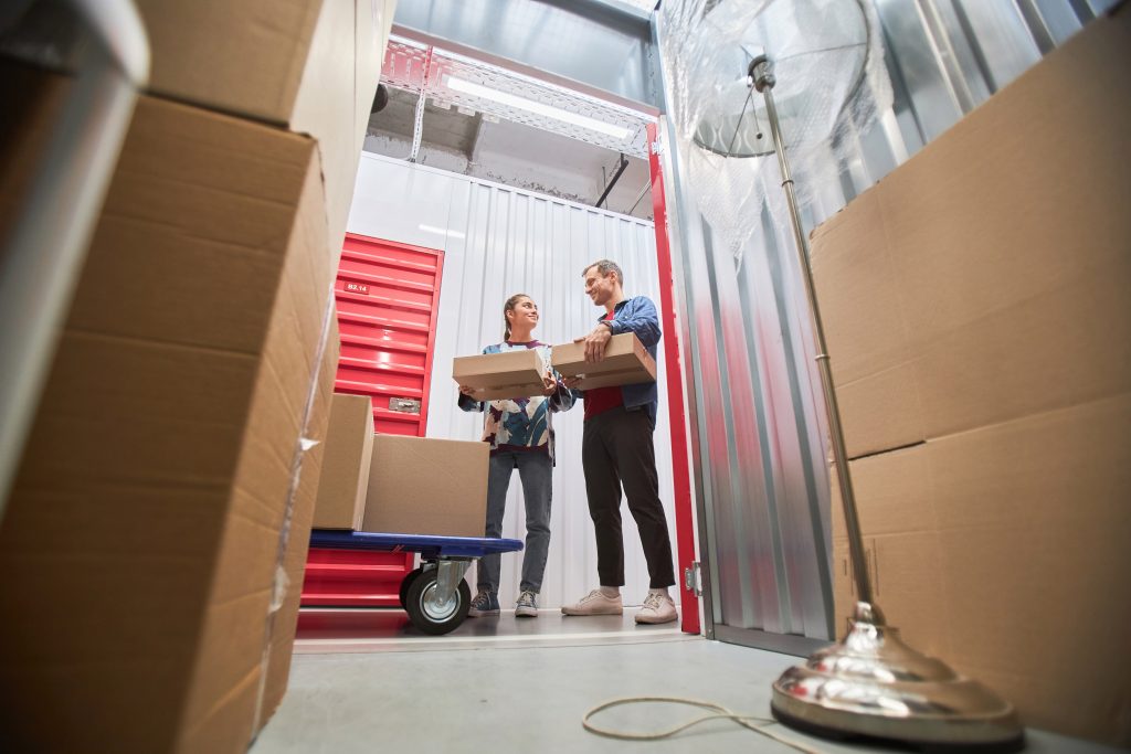 Two people standing in front of an open self-storage unit, loading boxes inside.