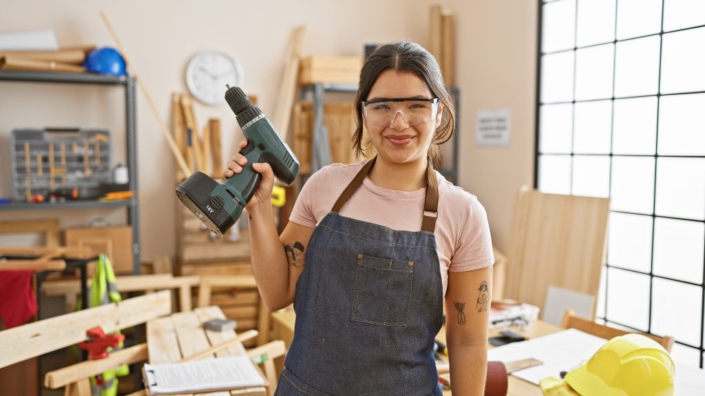 A smiling woman wearing safety goggles and holding an electric drill.