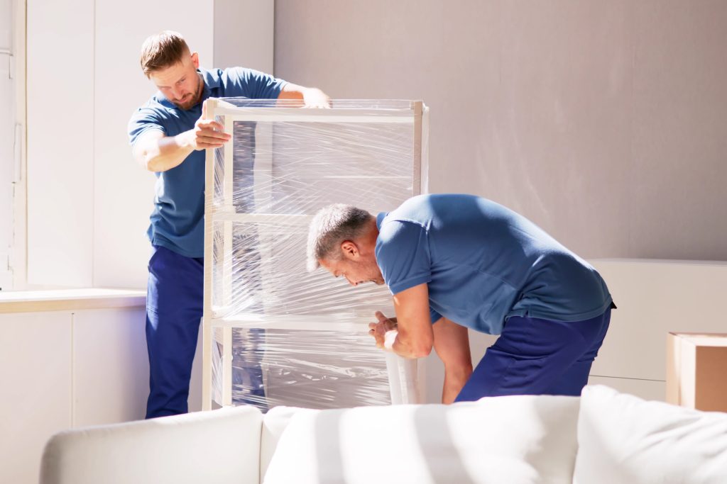 Two men wrap furniture in protective plastic wrap during home renovations.