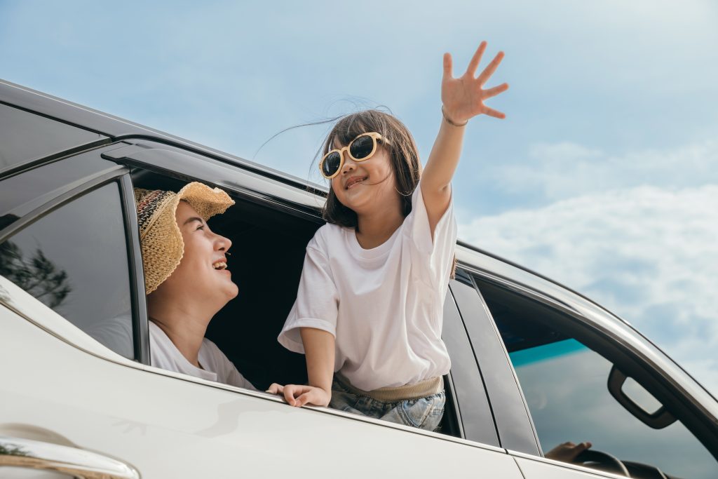 A young girl leaning out of an open SUV window, waving happily, while her mother smiles up at her from inside the vehicle.