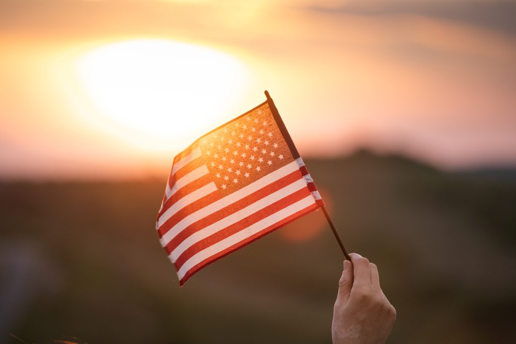 A hand holding up a small American Flag in a sunset.