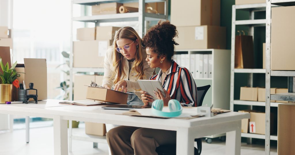 Two women in an inventory stock room of a small business, looking at a tablet on a desk.