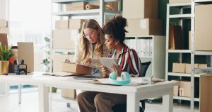 Two women in an inventory stock room of a small business, looking at a tablet on a desk.