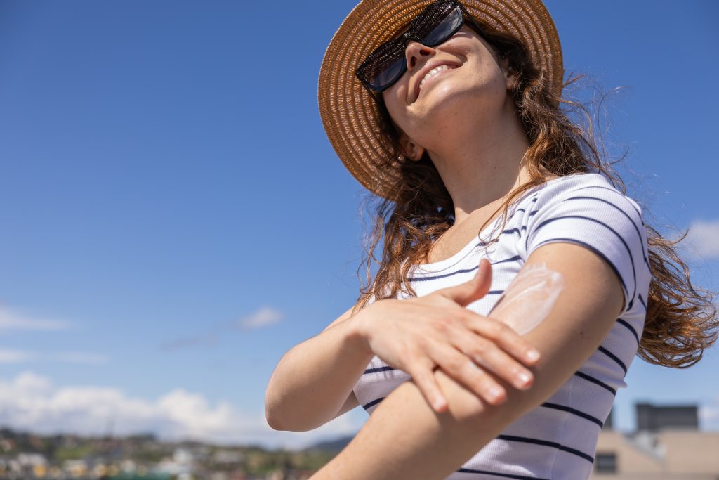 A woman in a hat and sunglasses applying sunscreen to her arm.
