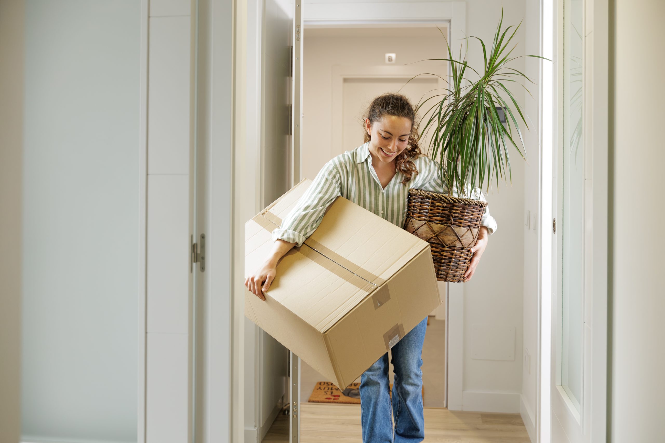Woman carrying a moving box and a houseplant into a new home.