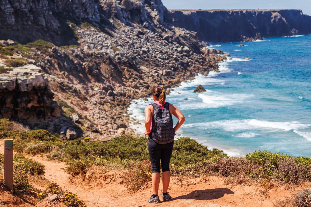 A woman standing on a hiking trail near a cliff overlooking the ocean.