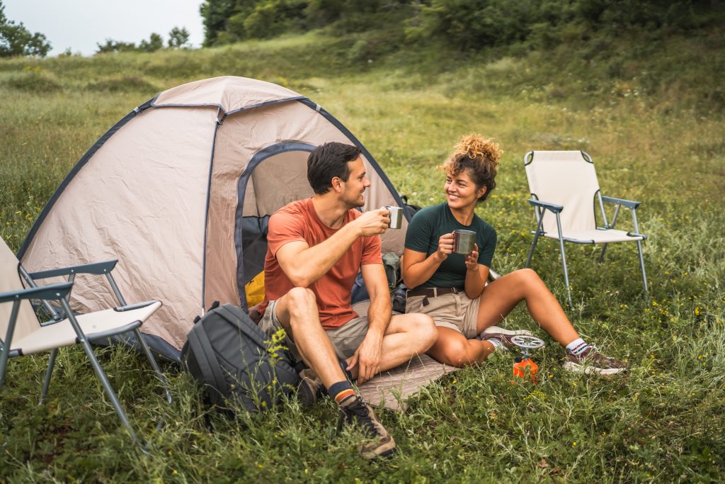 A couple sitting in a grassy field outside a camping tent and chairs.