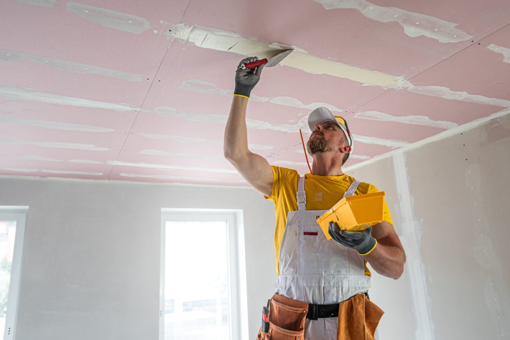 A man in overalls and safety goggles putting spackle on a ceiling.