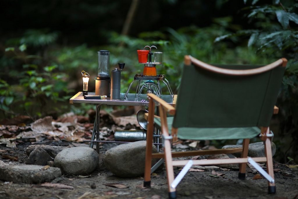 A camping chair, small table, candle, and coffee maker in the forest.
