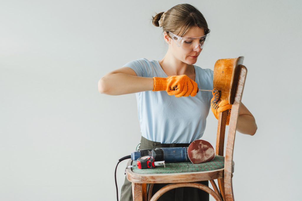 A woman in gloves and safety goggles fixing a chair, with power tools nearby.