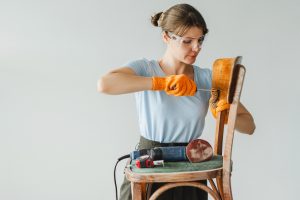 A woman in gloves and safety goggles fixing a chair, with power tools nearby.