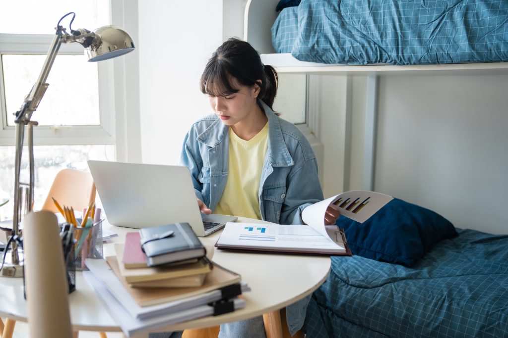 A female college student studying with a textbook and laptop at a desk in her dorm.