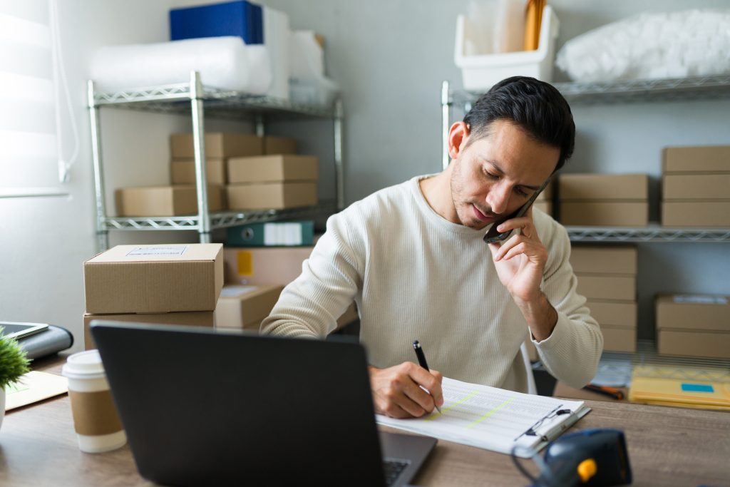A man in an inventory stock room talking on the phone and writing down an order.