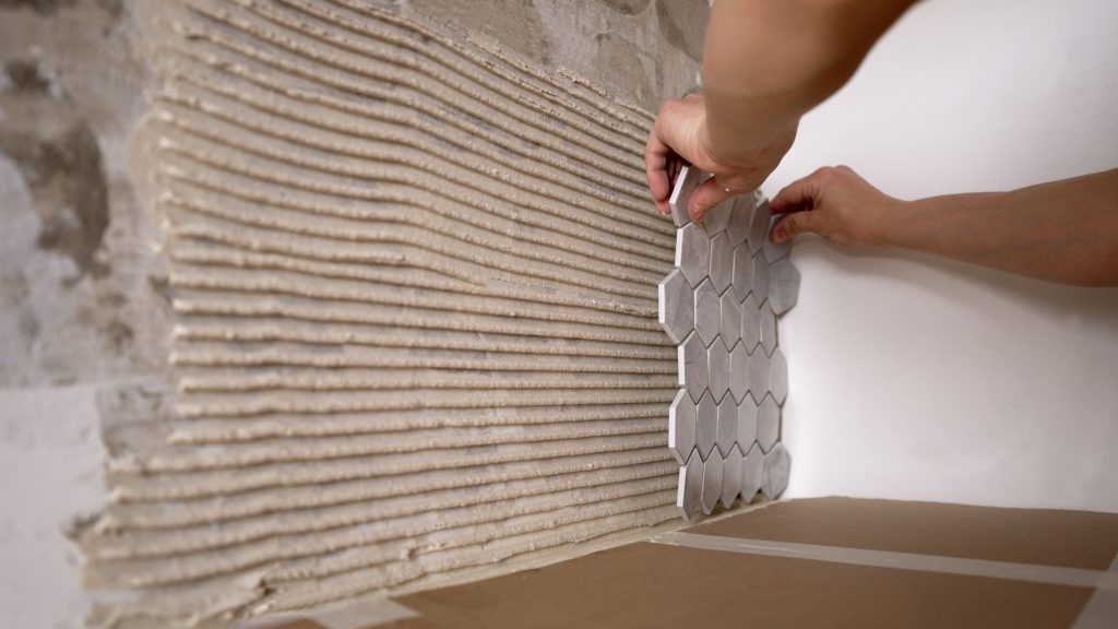 Two hands installing DIY kitchen backsplash tiles onto a wall with grout.
