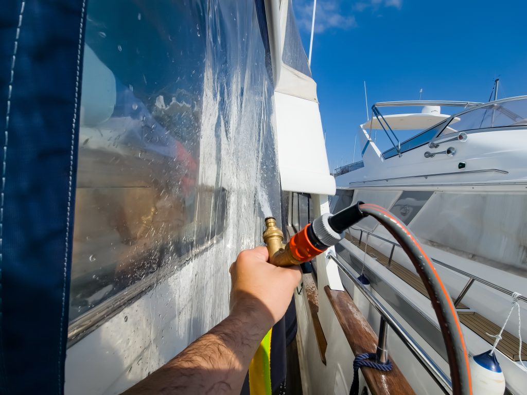 A hand holding a water hose spraying the side of a boat.
